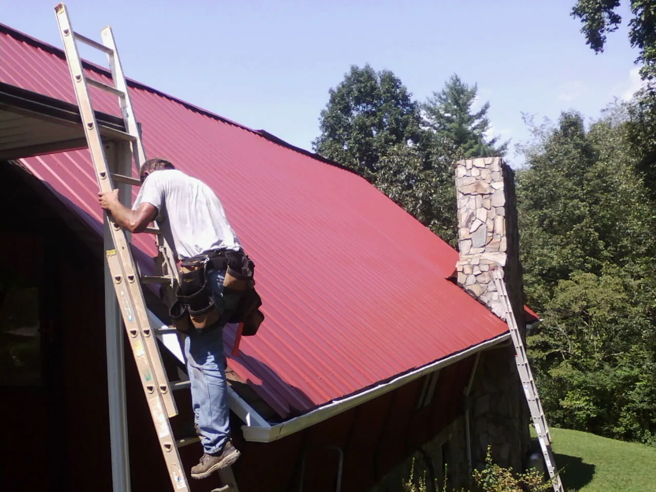 Roofer on a ladder beside a red metal roof and stone chimney — Soffit & Fascia Repair by Roofing Brothers in Gowen